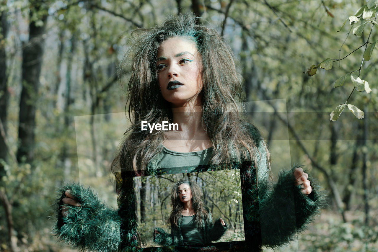 Serious young woman with spooky make-up holding picture frame while standing in forest