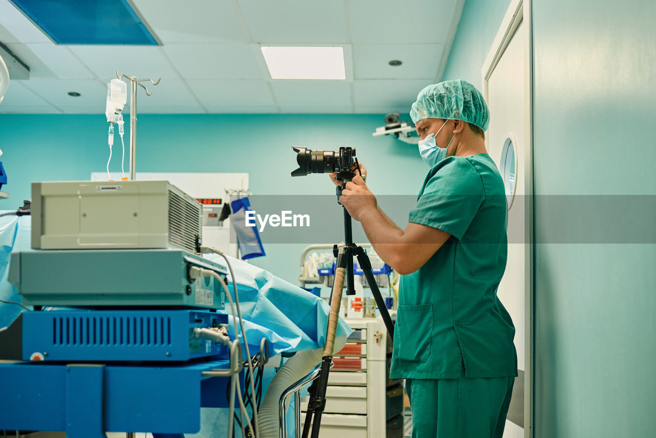 Side view of unrecognizable young male medical photographer in sterile mask and uniform taking pictures on professional camera during surgery in modern operating room