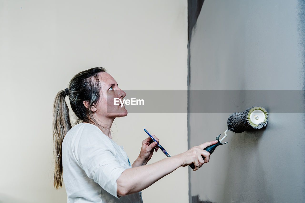 Woman catching with a hand a paint roller full of grey painting and with the other a brush. she is upping and downing the roller covering the wall with grey painting what remains wet. horizon