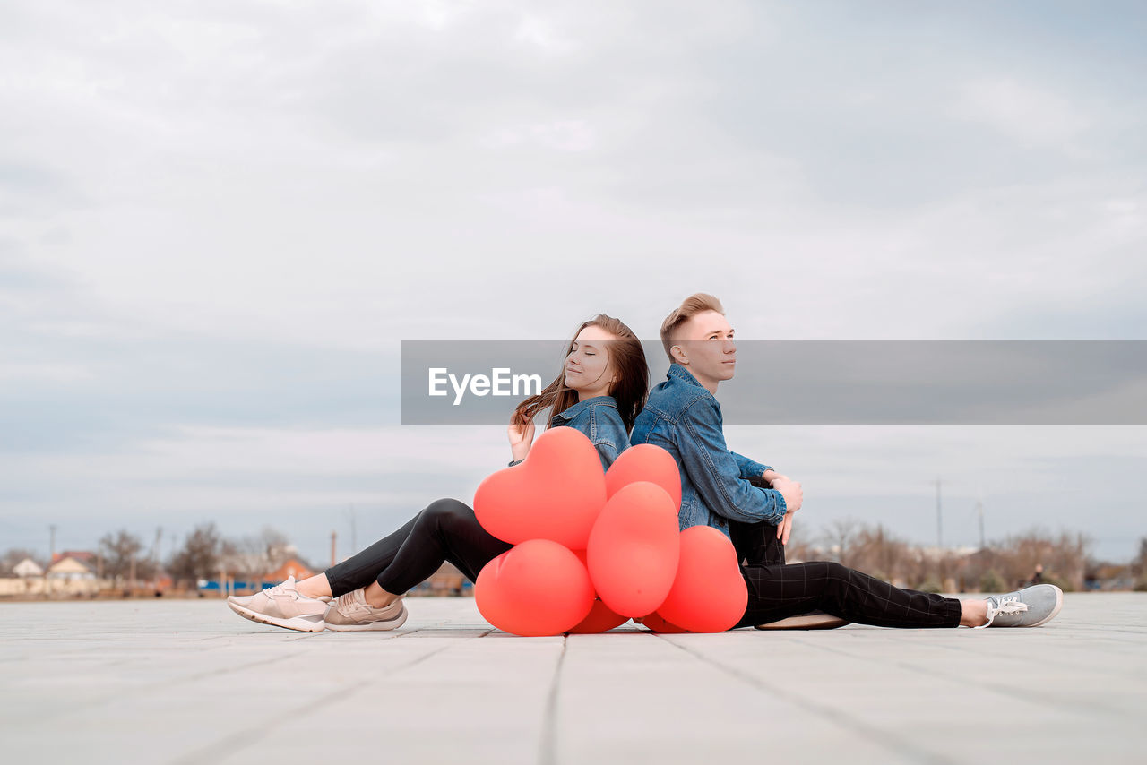 Young couple sitting back to back in the street holding a pile of red balloons spending time