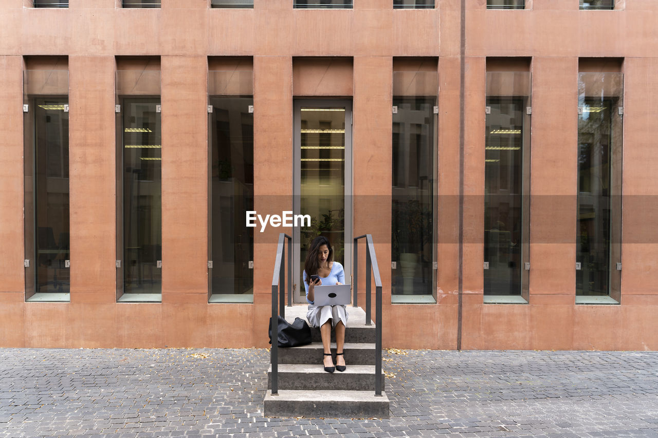 Businesswoman using laptop while sitting on steps at entrance of office building