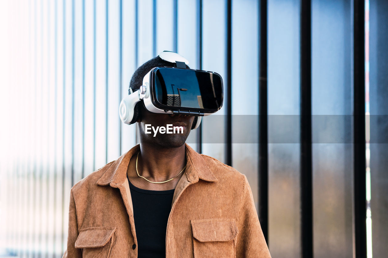 African american male in vr goggles exploring cyberspace while standing on street near modern building with metal wall in city
