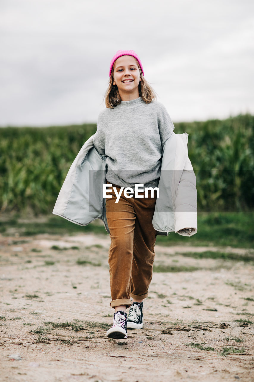 Cheerful beautiful teenage girl in yellow raincoat and pink hat confidently walks in the corn field