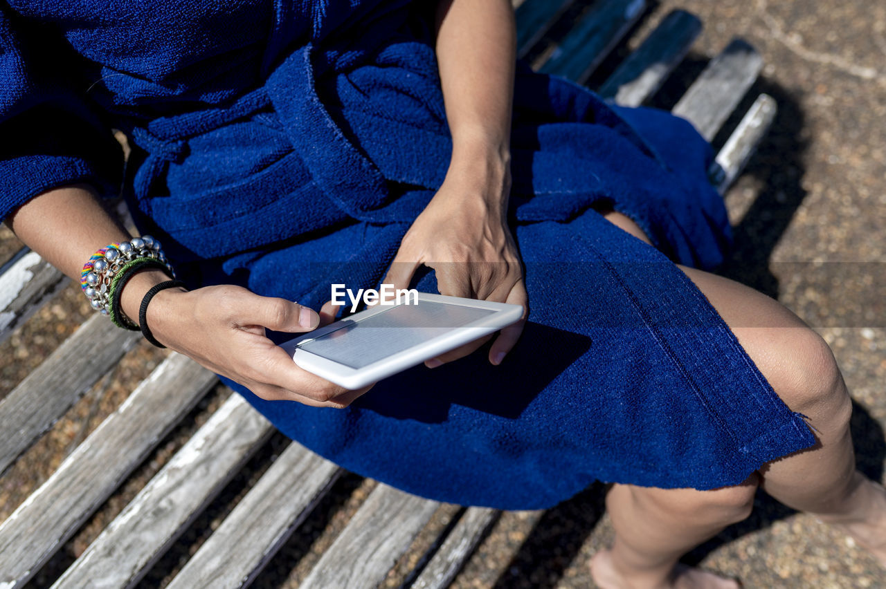 Close-up view of woman in robe reading an e-book sitting on a bench outdoors.