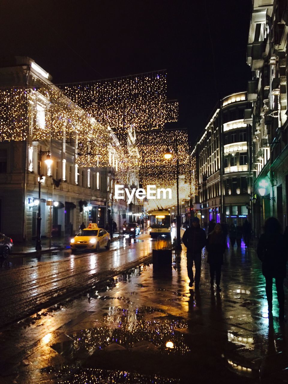 People walking on wet sidewalk by illuminated buildings at night