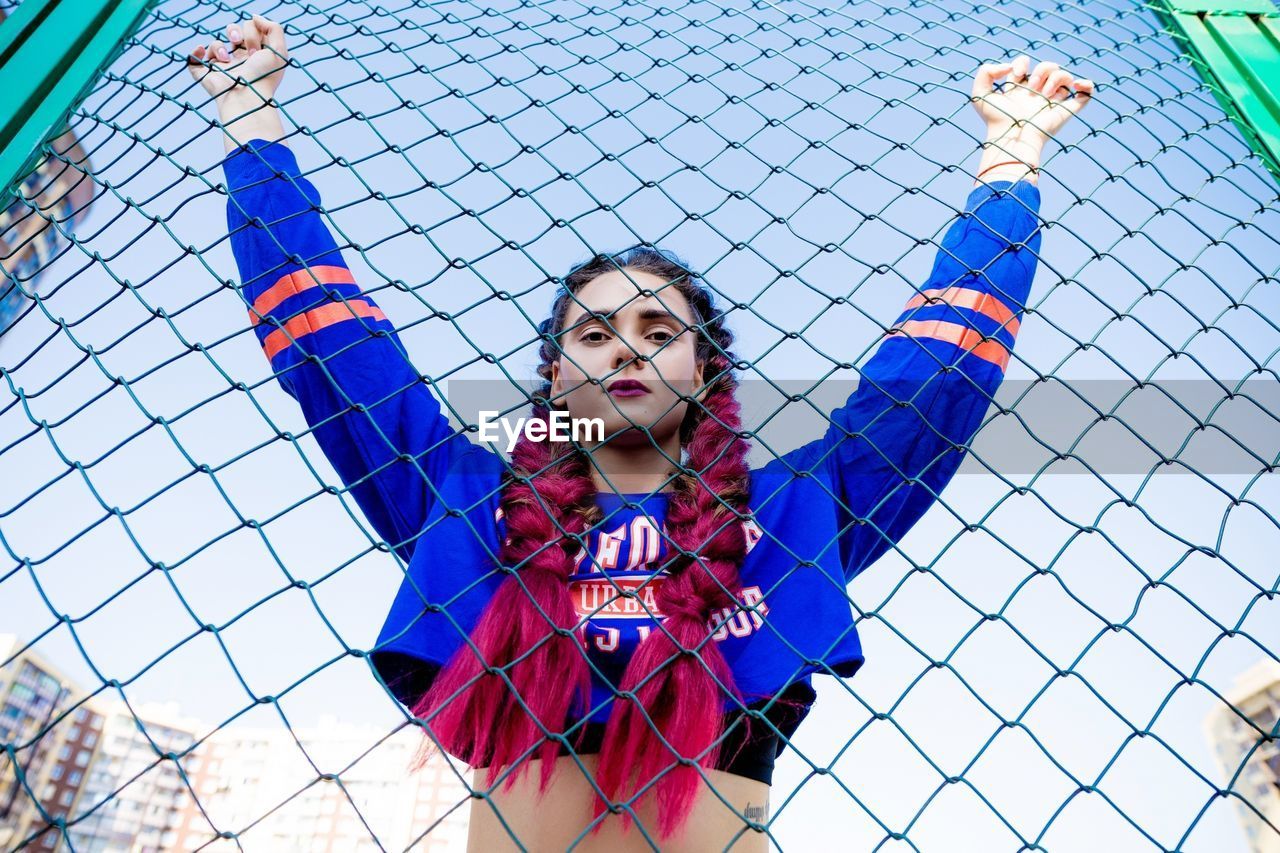 Portrait of young woman with arms raised standing by chainlink fence against sky