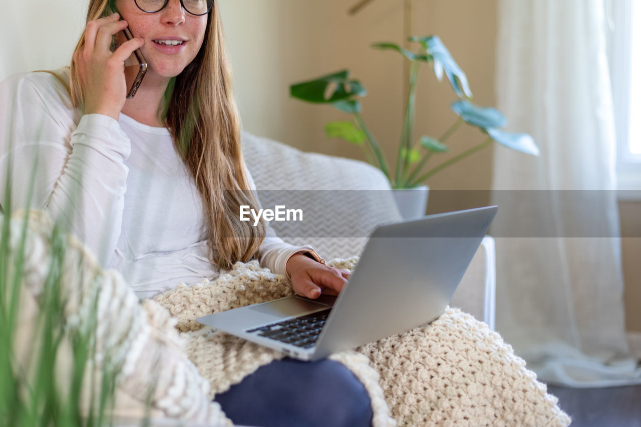 Young woman at home on the couch using laptop and cellphone