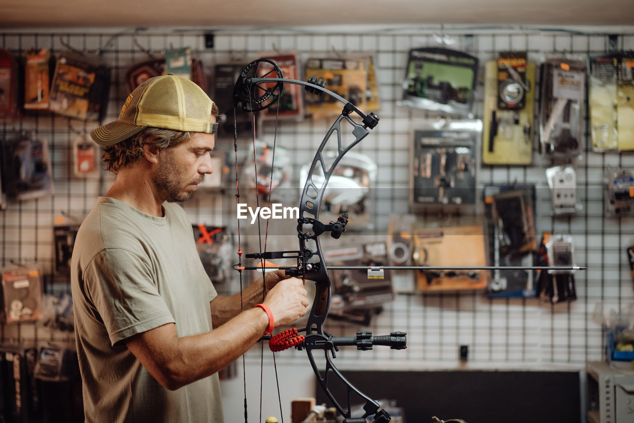 Side view of busy male hunter adjusting compound bow with arrow while standing in garage and preparing for shooting