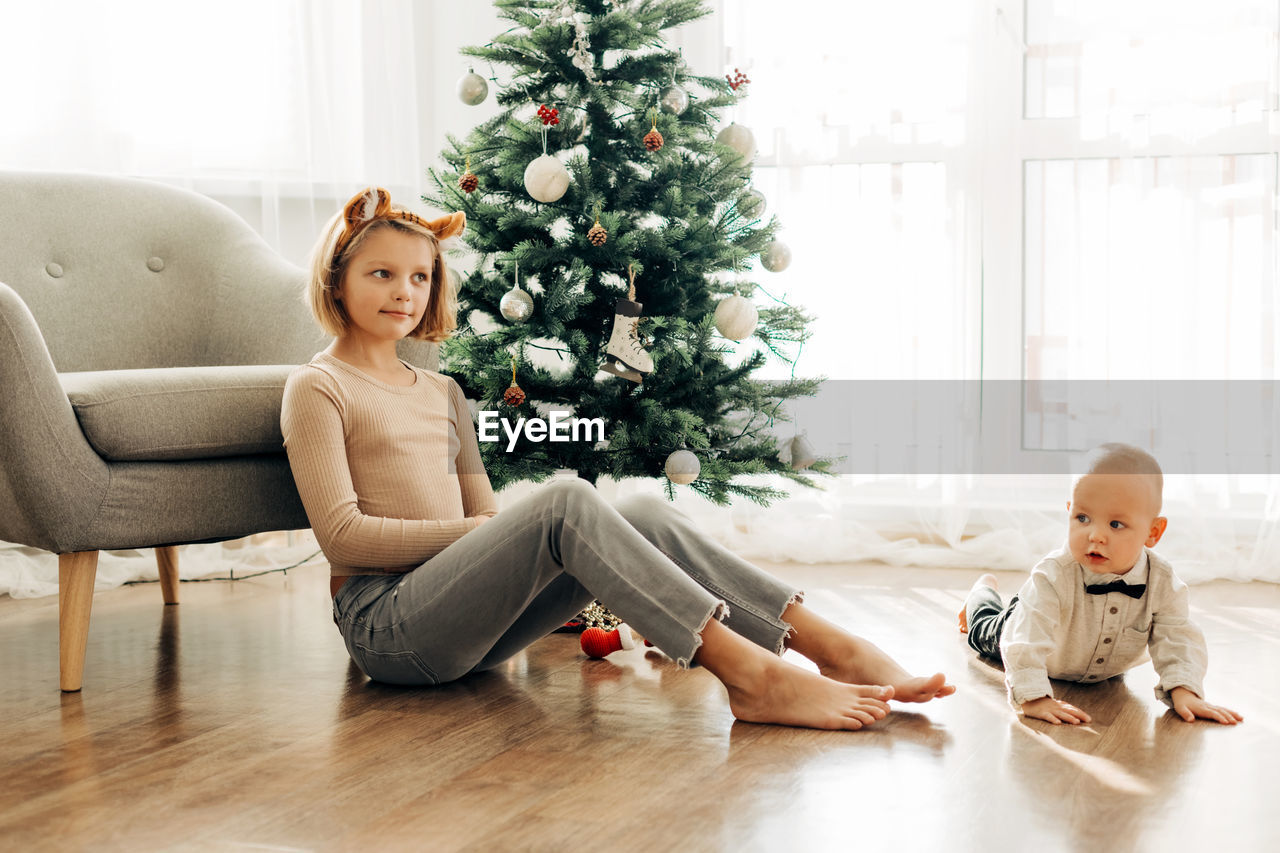 A charming girl and her younger brother play together on the floor next to the christmas tree 