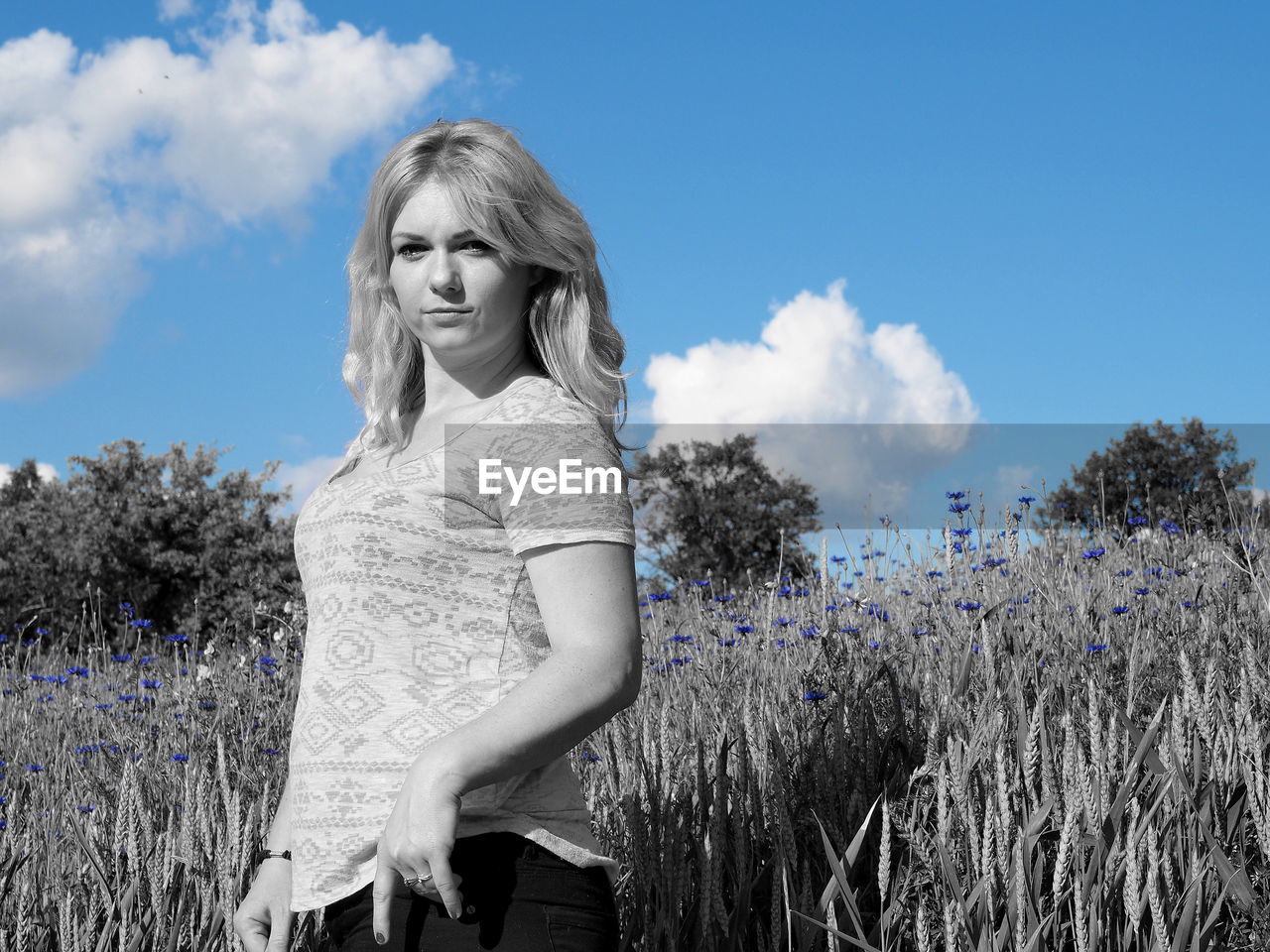 PORTRAIT OF SMILING YOUNG WOMAN STANDING AGAINST TREES ON FIELD