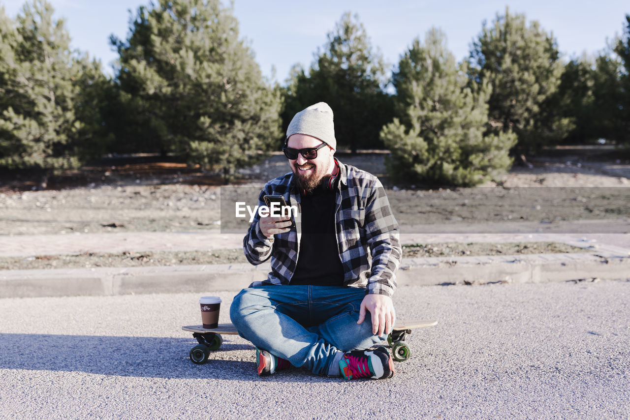 Modern young man sitting on a longboard looking at his mobile phone