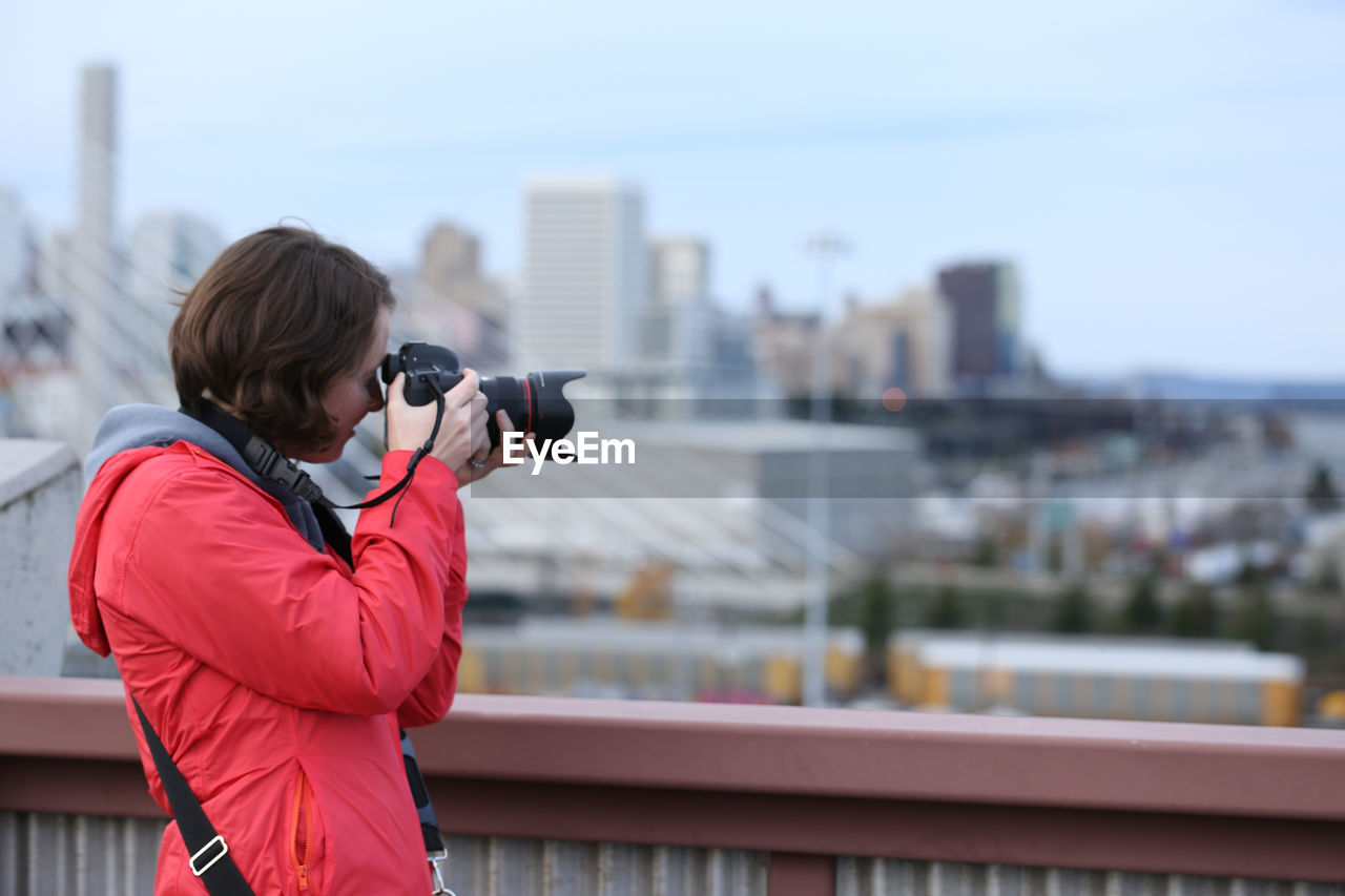 Side view of woman photographing in city against sky during winter