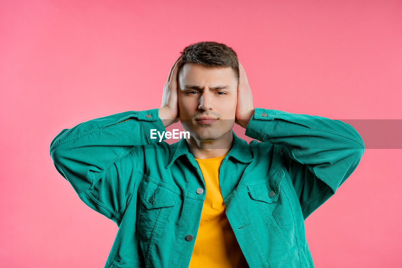 colored background, studio shot, one person, hood, outerwear, young adult, clothing, adult, jacket, portrait, indoors, front view, men, sleeve, waist up, photo shoot, hoodie, green, pink, emotion, person, standing, casual clothing, looking, serious, copy space, pink background, eyes closed, lifestyles, brown hair, blue