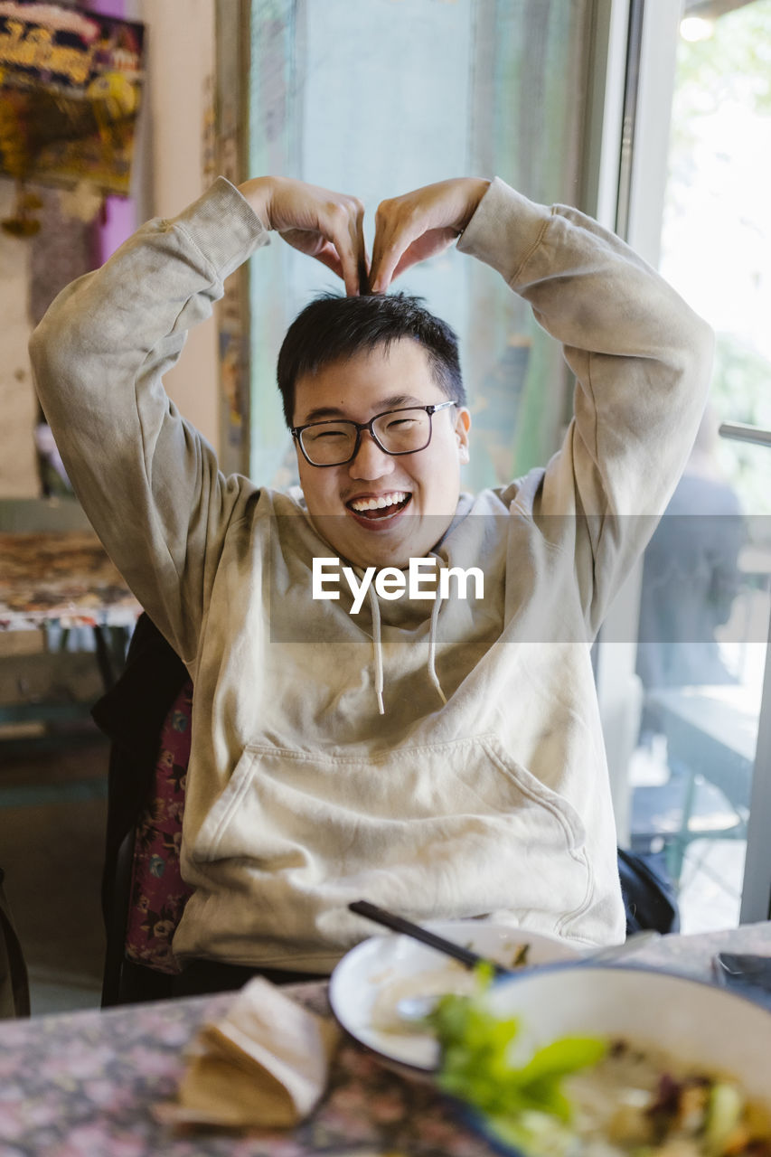 Happy young man making heart shape with hands while sitting at restaurant