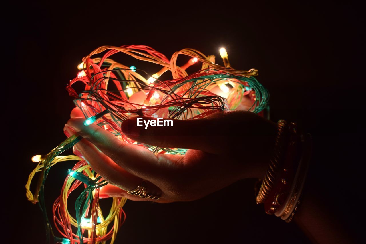 Cropped hands of woman holding illuminated string lights against black background