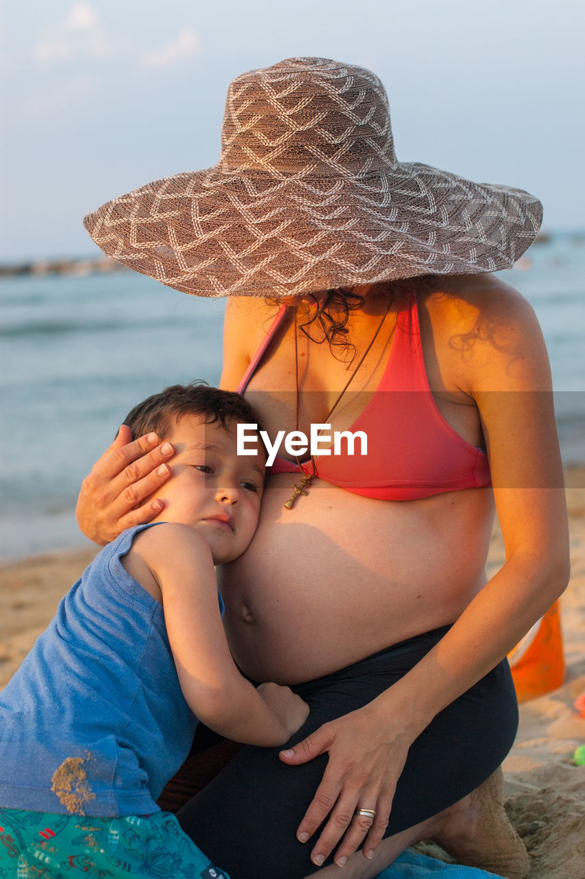 Son touching stomach of pregnant woman while sitting at beach against sky