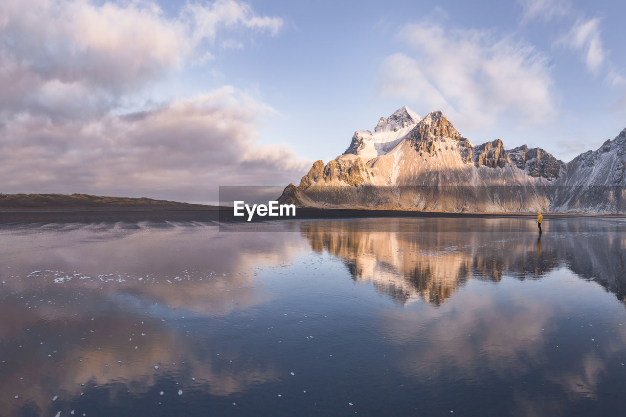 Anonymous tourist standing on wet shore near calm sea and rough mountains during majestic colorful sunset