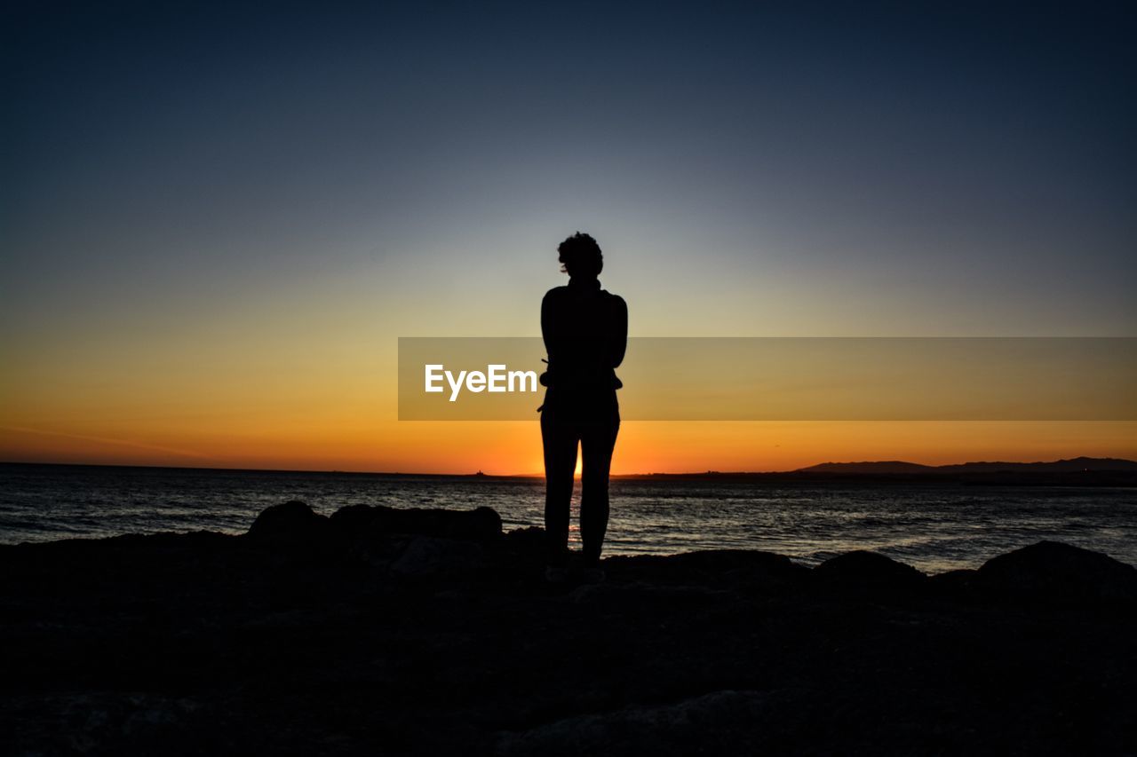 SILHOUETTE MAN STANDING ON ROCK AT BEACH AGAINST SKY