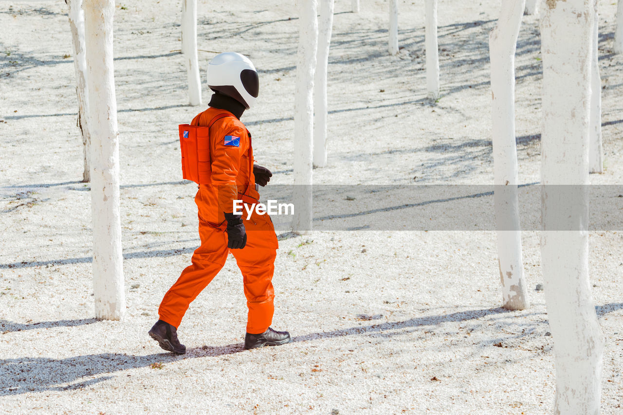 Side view of unrecognizable male cosmonaut in bright orange spacesuit and helmet walking on sandy ground amidst white trees on sunny day on new planet