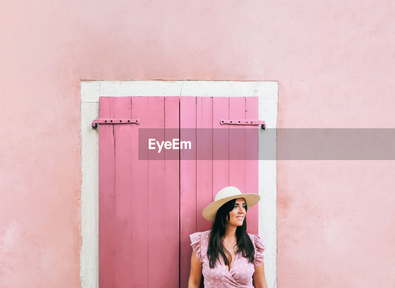 Beautiful young woman in pink dress standing in front of pink wall and wooden window shutter.