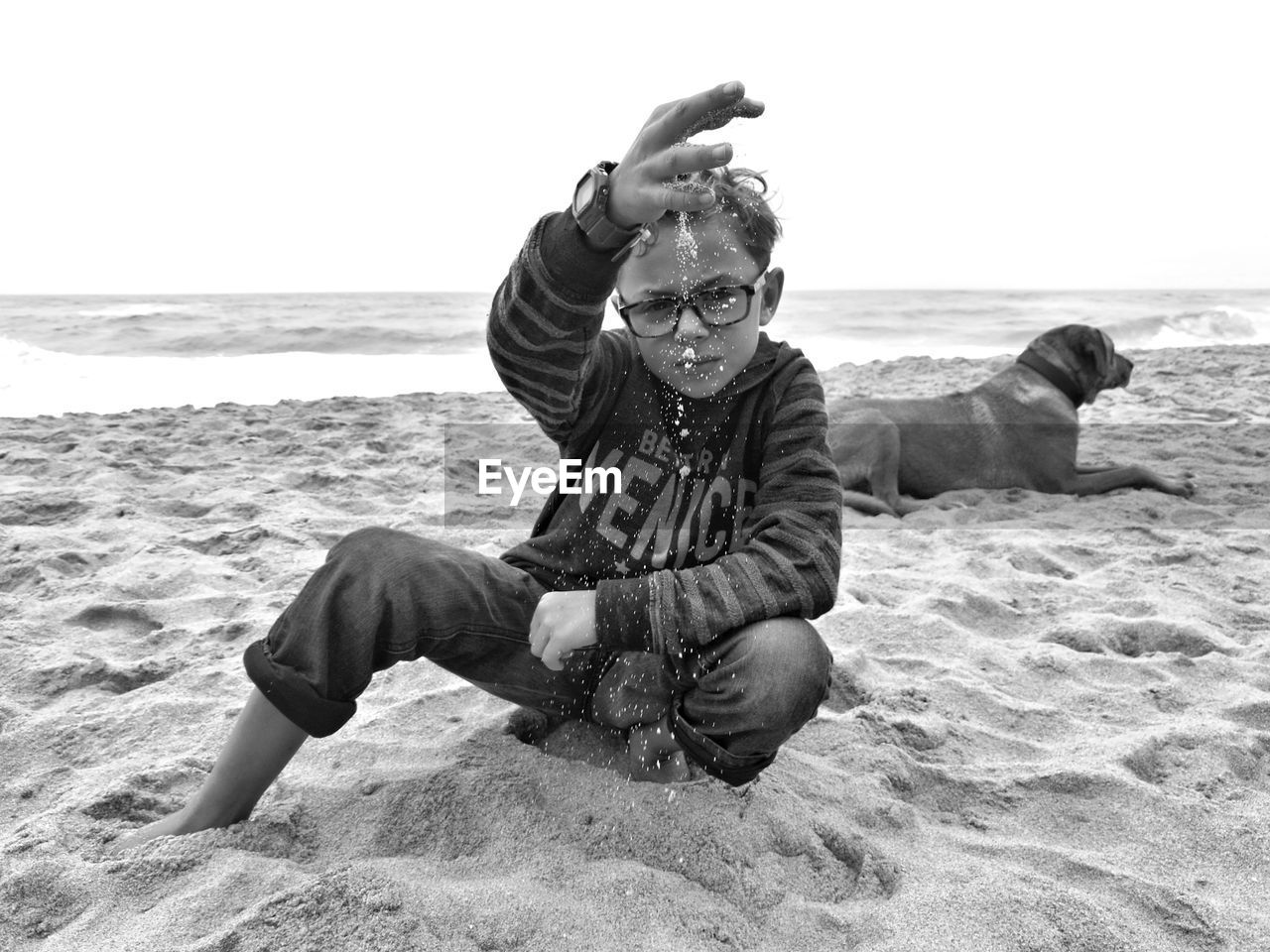 Boy playing with sand by dog at beach