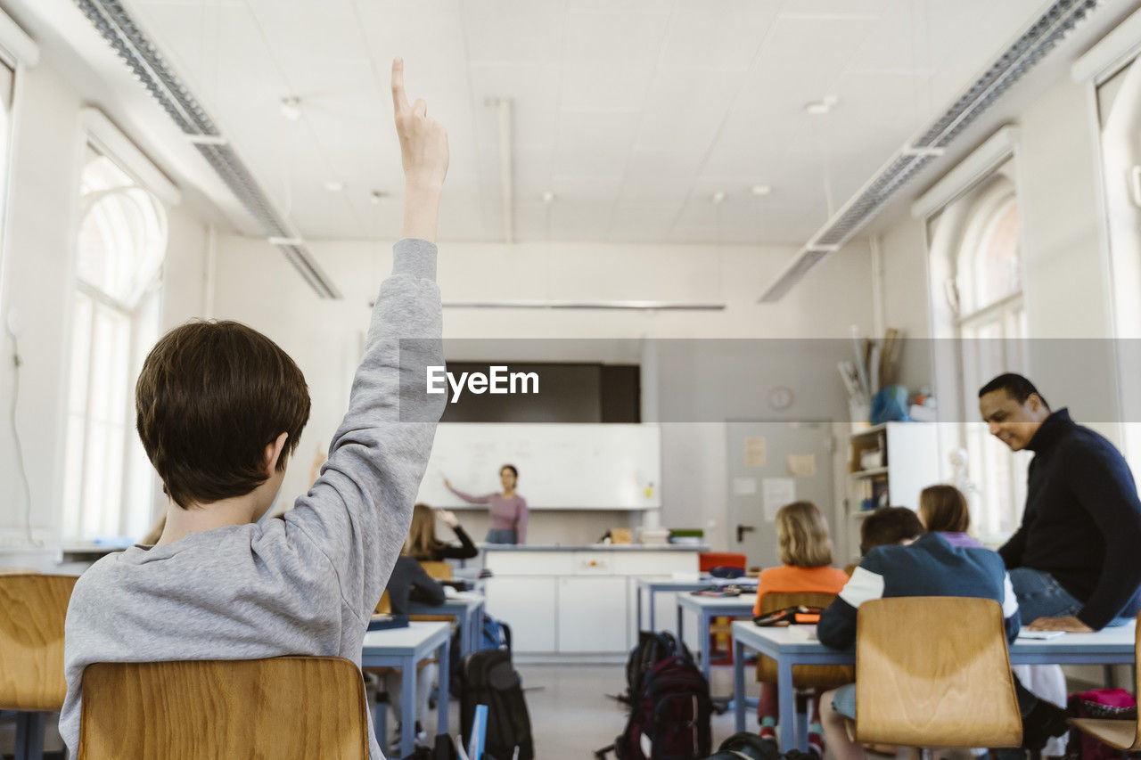 Rear view of schoolboy raising hand while attending lecture in classroom