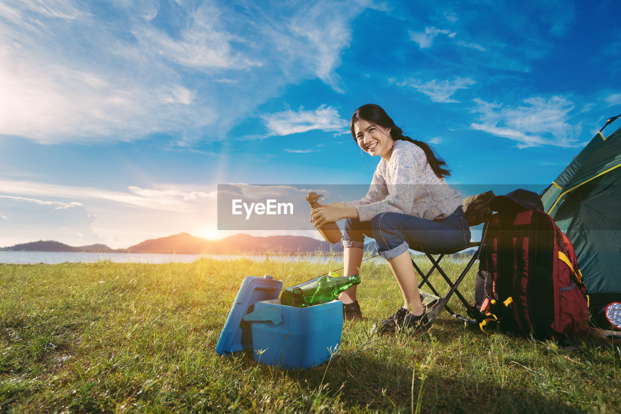 Portrait of smiling young woman camping on grassy field against blue sky