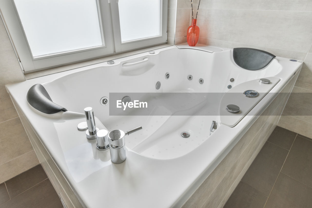 High angle of large white bathtub with hydromassage supplies located near window in modern luxurious bathroom at home