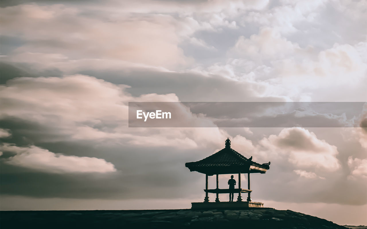 Silhouette person standing in gazebo against cloudy sky