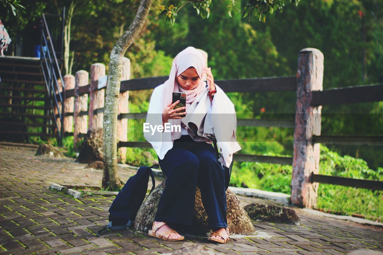Woman wearing hijab taking selfie with smart phone while sitting in park