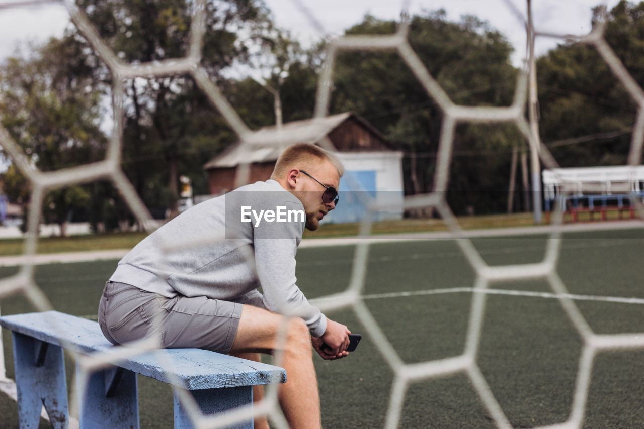 Side view of man sitting on bench seen through net