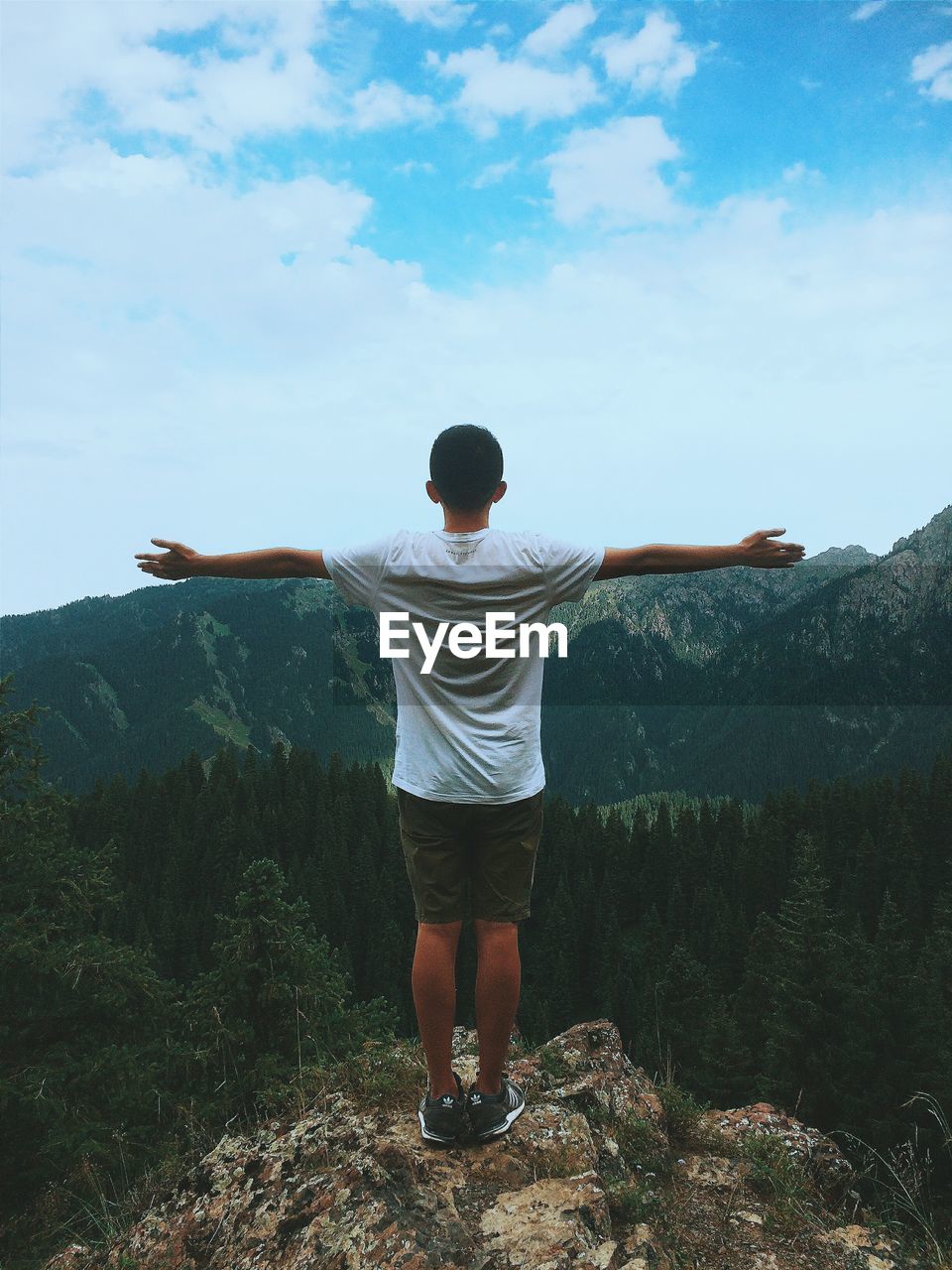 Rear view of young man standing with arms outstretched on rock against sky