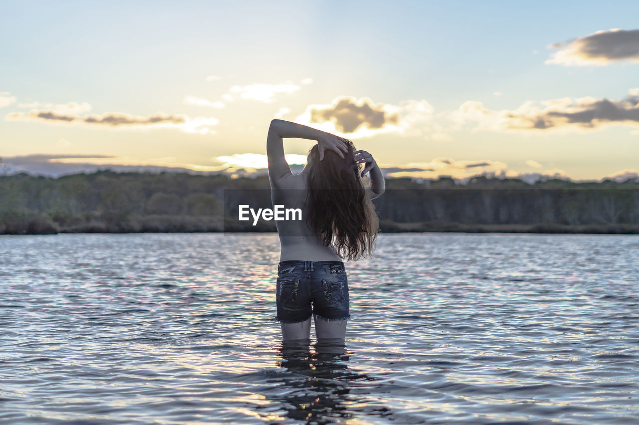 Rear view of shirtless woman standing in lake against sky