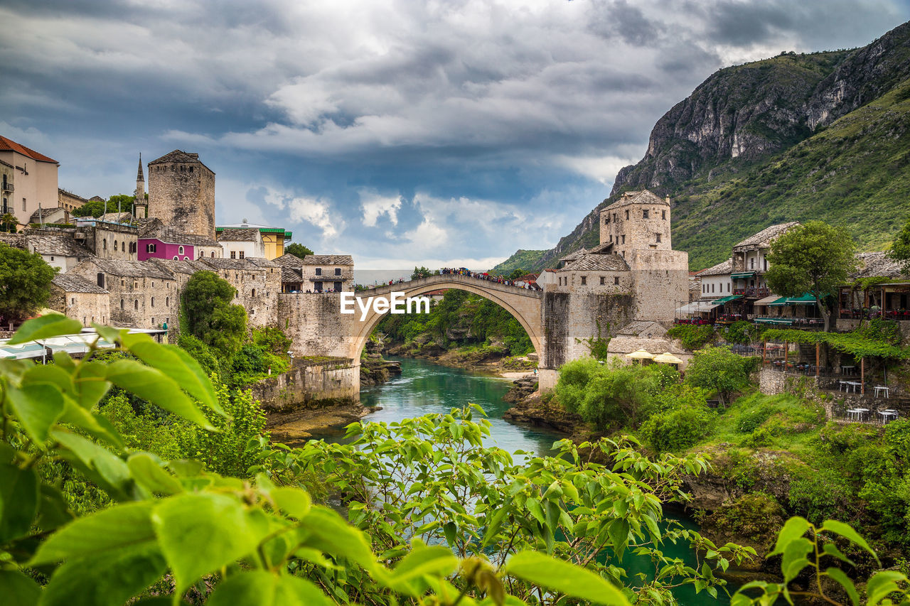 Arch bridge over river amidst buildings against cloudy sky