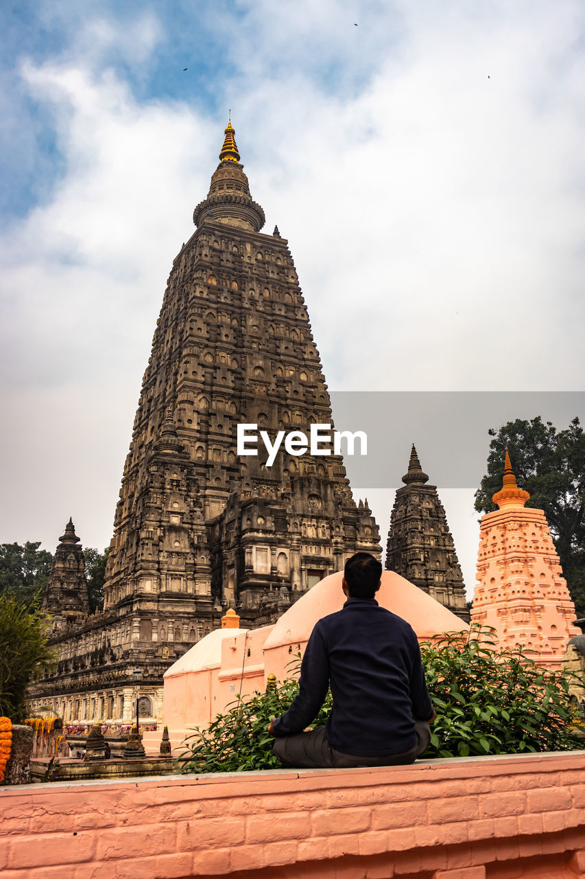 Rear view of man sitting outside temple against sky