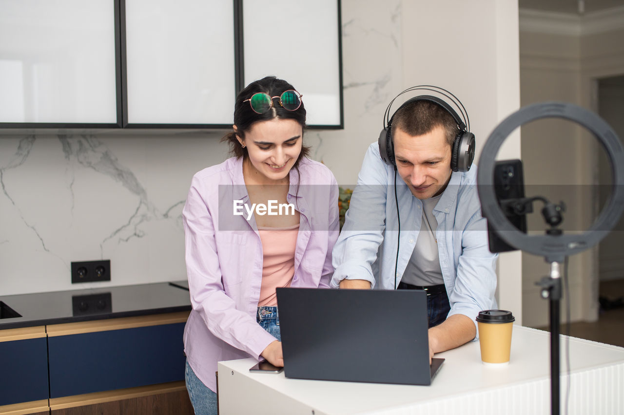 A man with headphones and a girl with glasses are streaming and looking at a laptop in the room