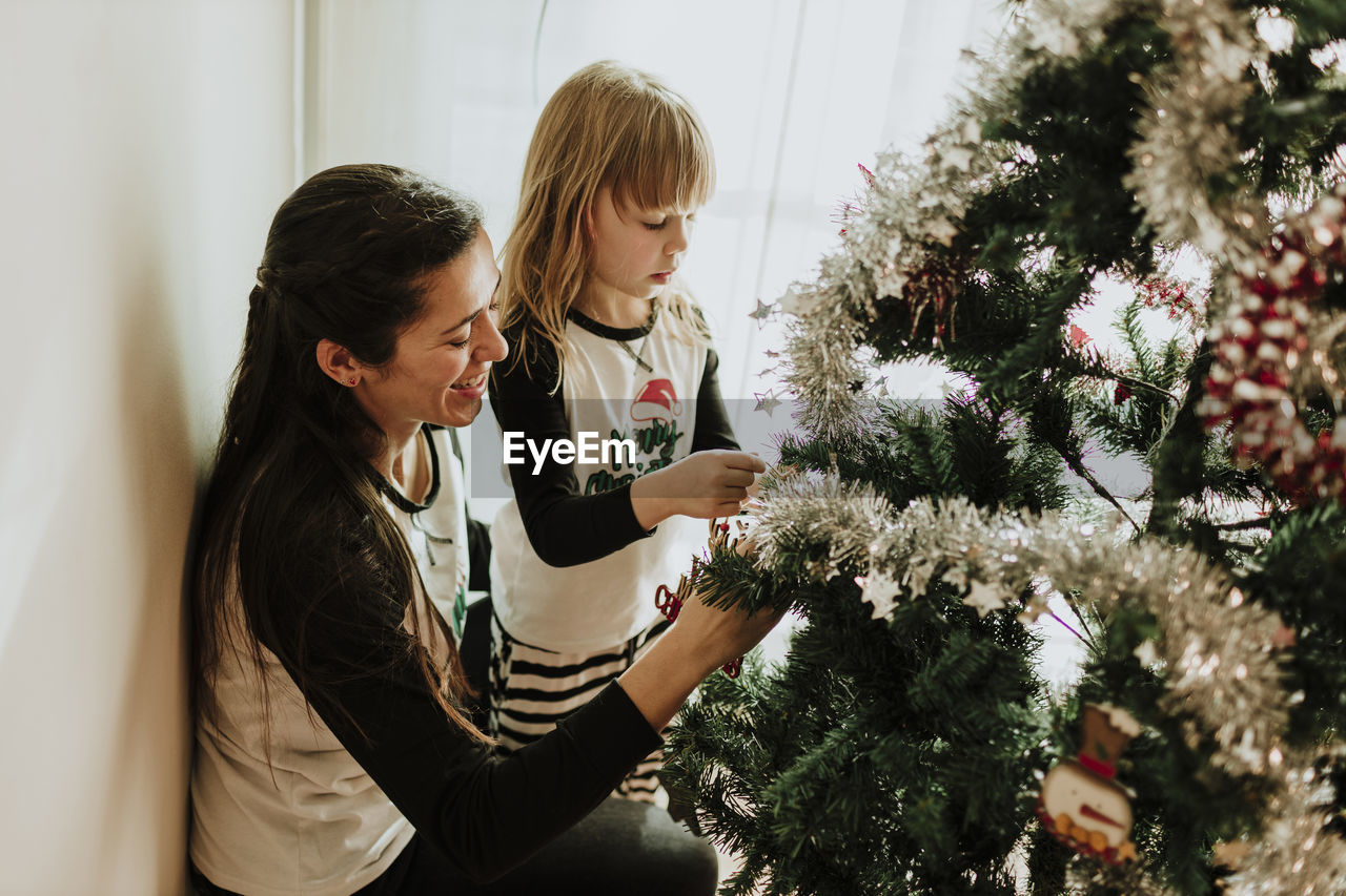 Mother with daughter decorating christmas tree against wall at home