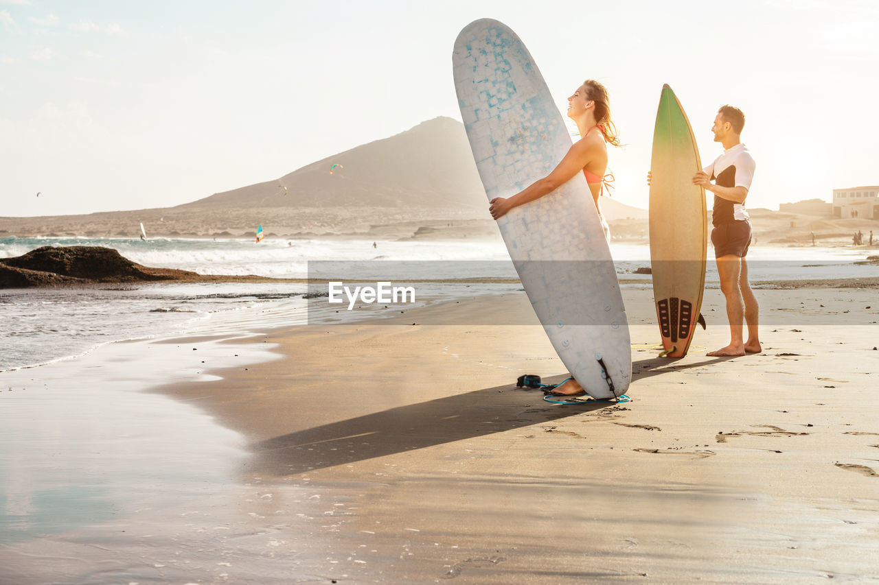 Couple standing with surfboard at beach against sky