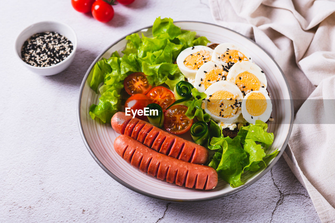 Plate with sausages, egg sandwich, tomatoes and lettuce leaves on the table