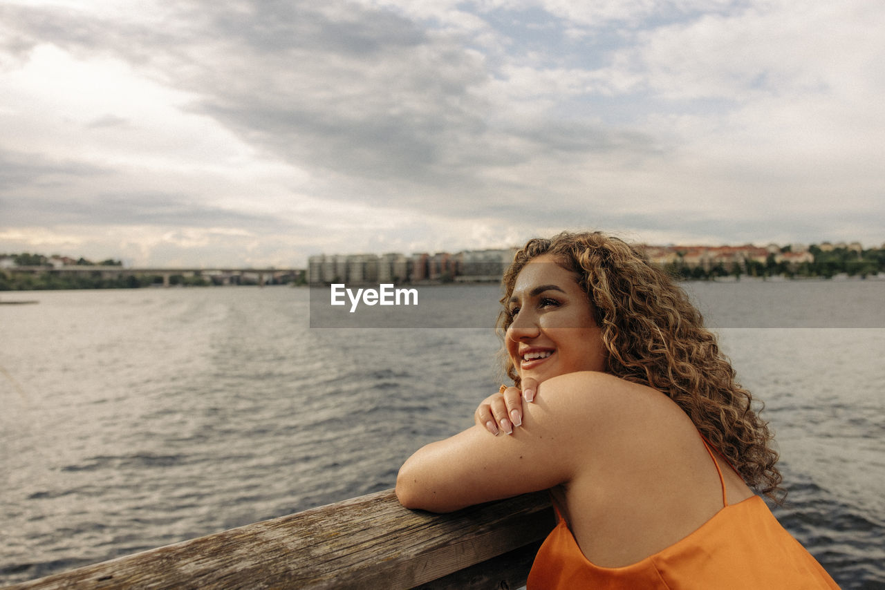 Happy woman with curly hair leaning on railing against sky