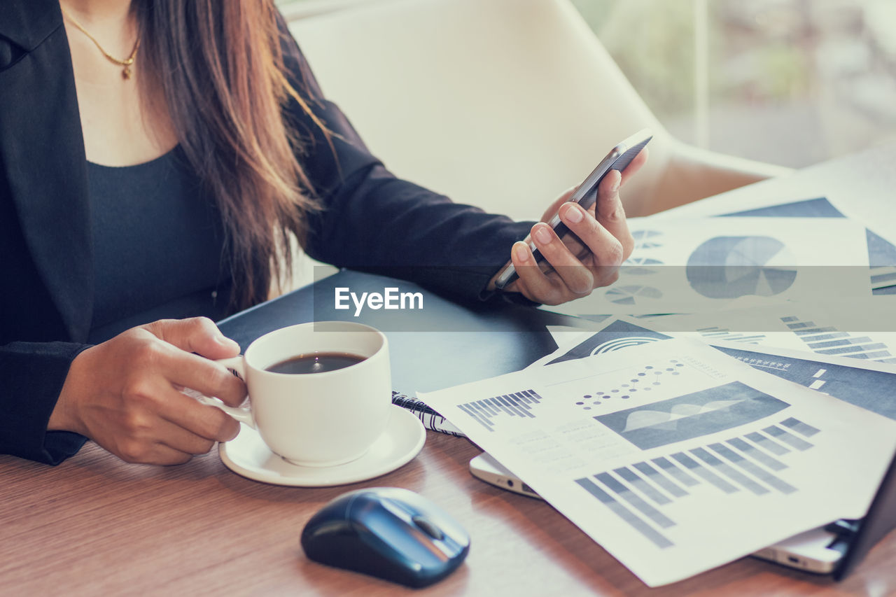 Midsection of woman with coffee using phone by laptop on table