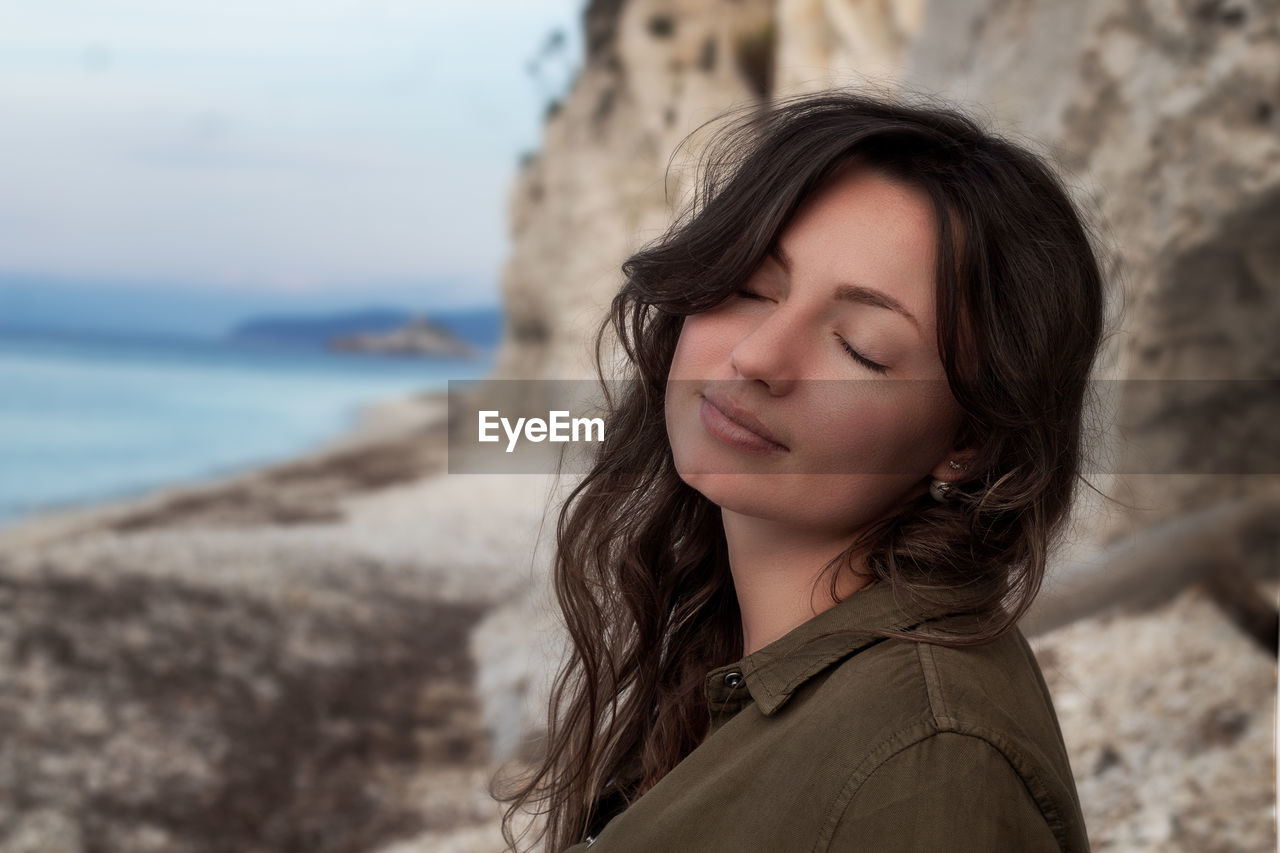 Side view of beautiful woman with eyes closed sitting against rock formation on beach