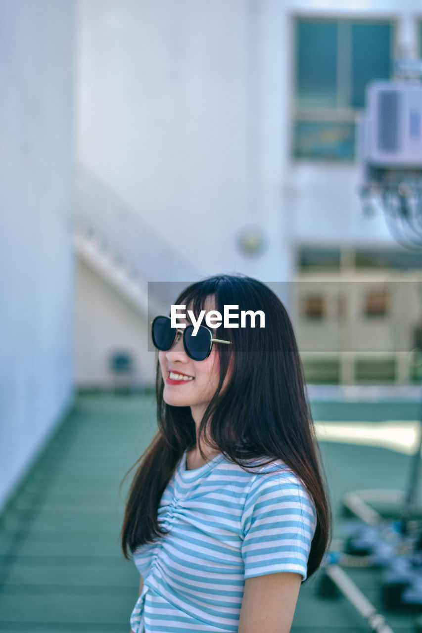 Cheerful young woman wearing sunglasses standing against building