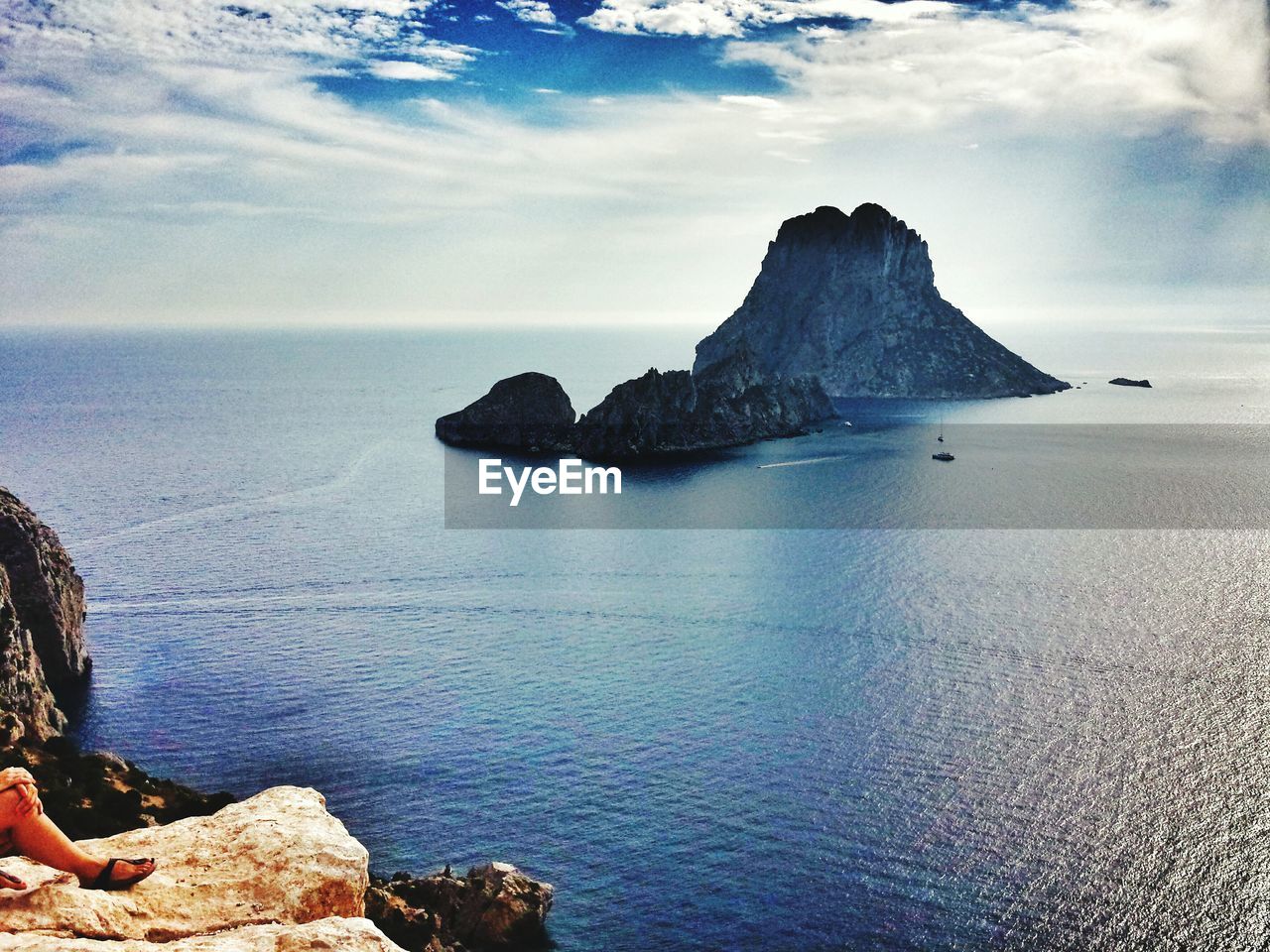 Low section of person sitting on cliff by sea against sky