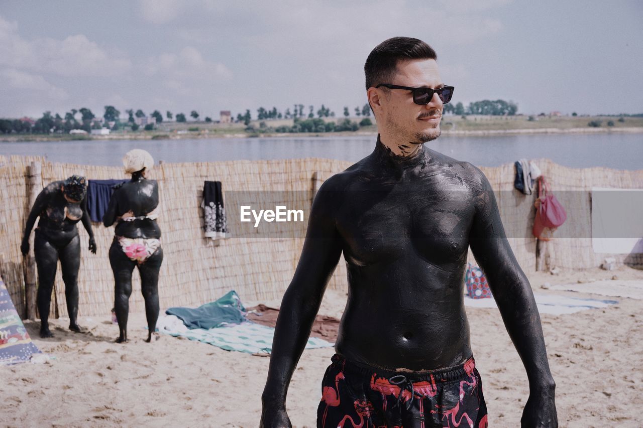 Man with black mud on body standing at beach against sky