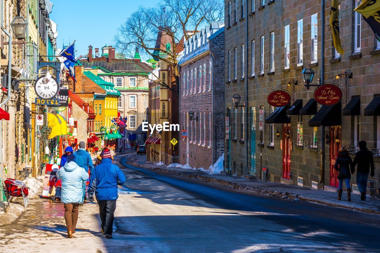 REAR VIEW OF PEOPLE WALKING ON STREET BY BUILDINGS