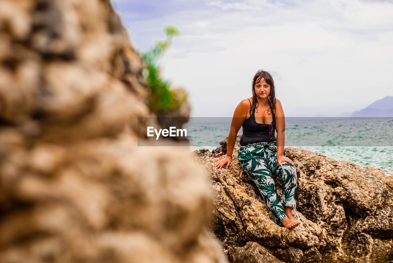 Portrait of woman sitting on rock at beach against sky