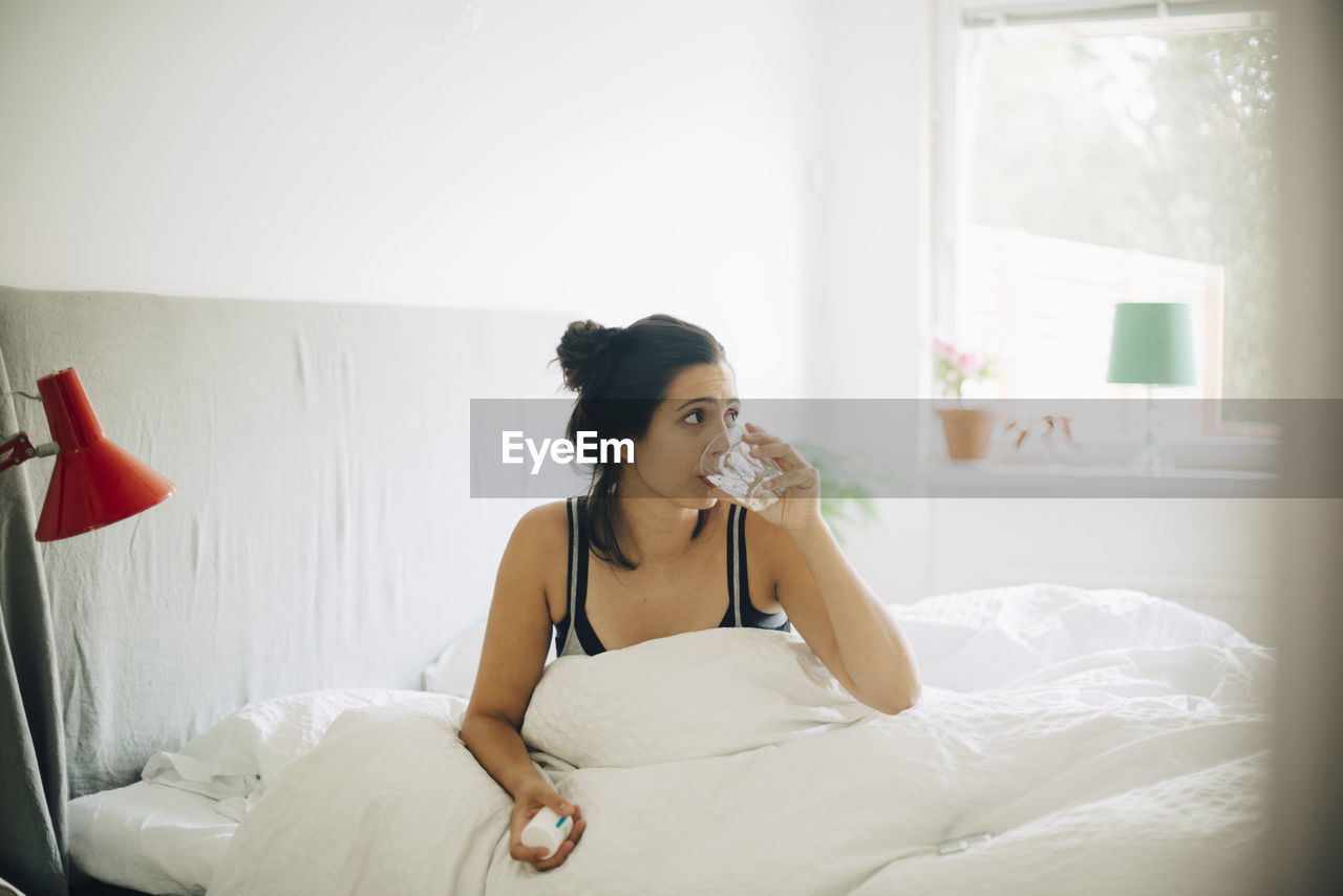 Woman drinking water while holding pills on bed at home