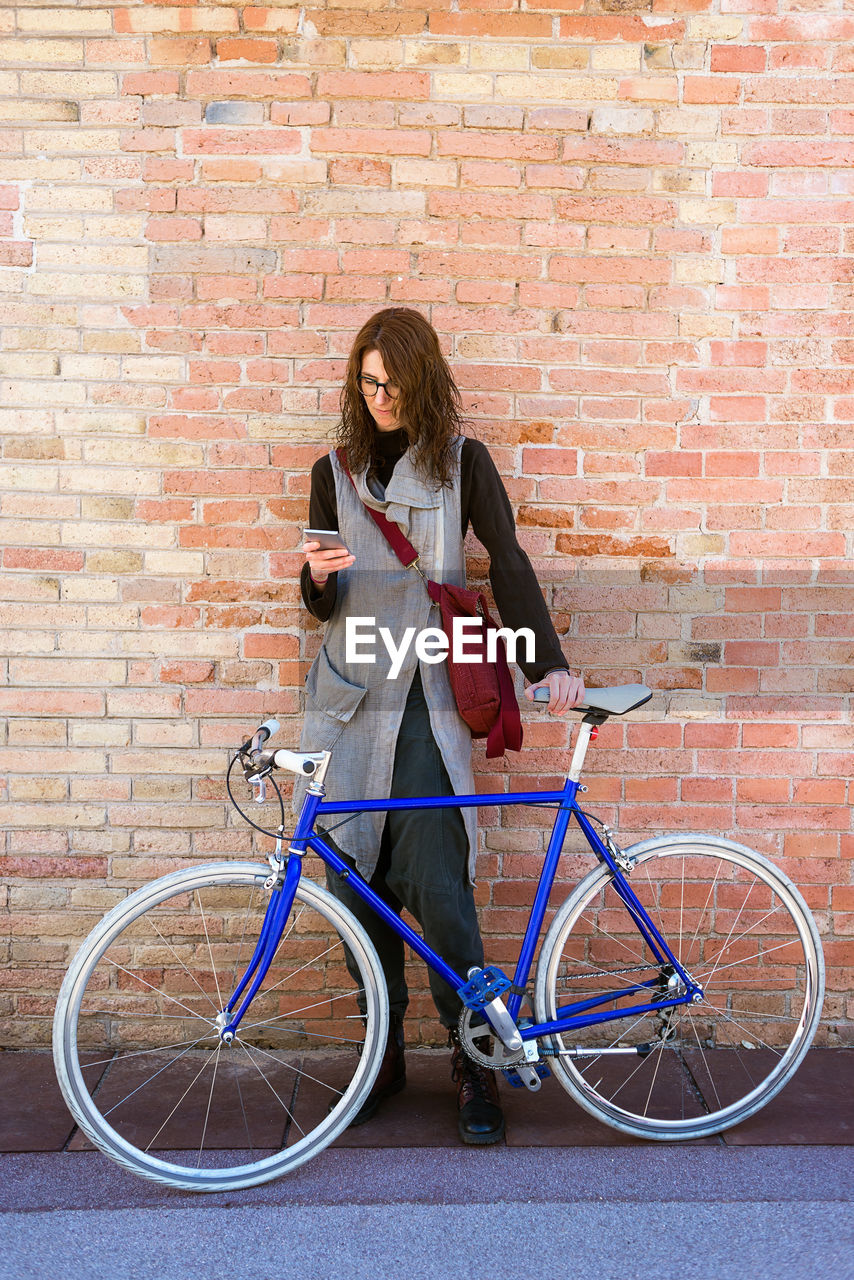 Smiling young woman with bike leaning on wall while using mobile