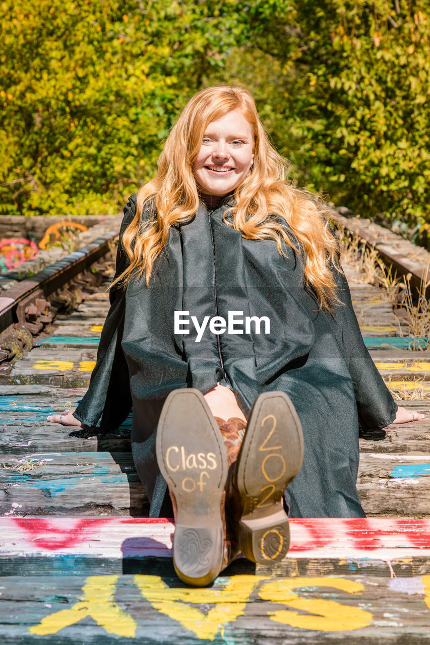Portrait of smiling girl in graduation gown sitting on wood with text and numbers on shoes